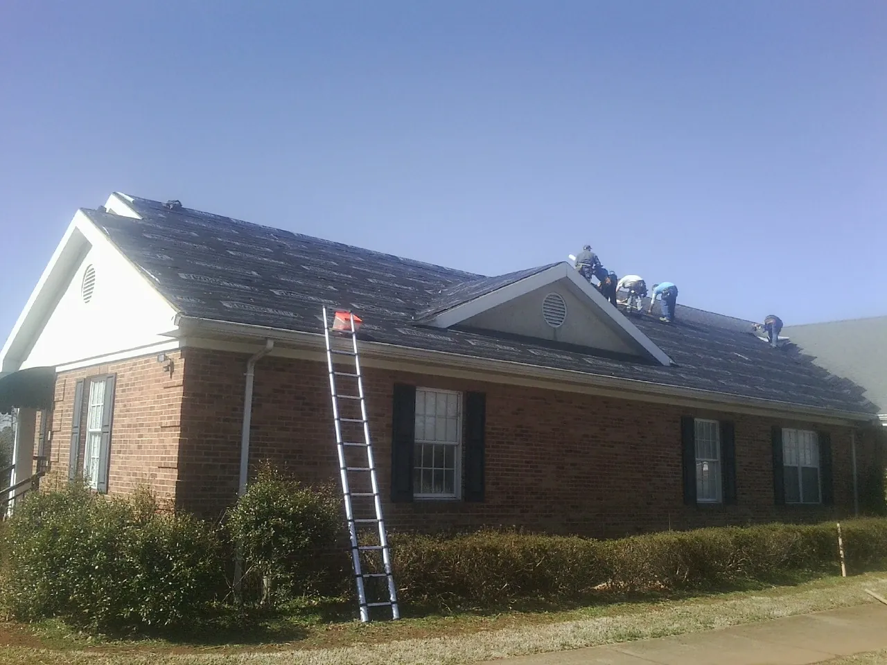 Crew tearing shingles off a brick ranch roof with a ladder set — Emergency Roof Repair work by our crew in Harrah